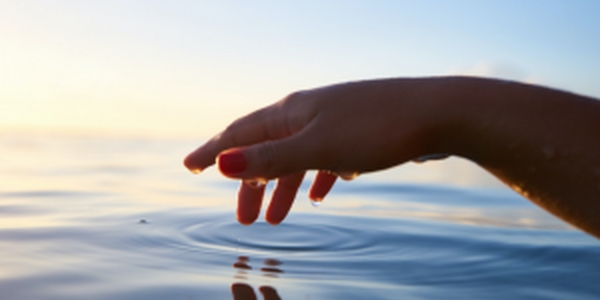 Hand trailing through clear blue water and causing a gentle ripple